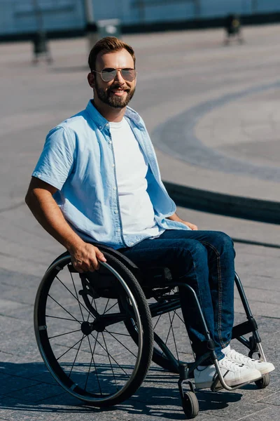 Retrato de hombre guapo en gafas de sol usando silla de ruedas en la calle y mirando a la cámara - foto de stock