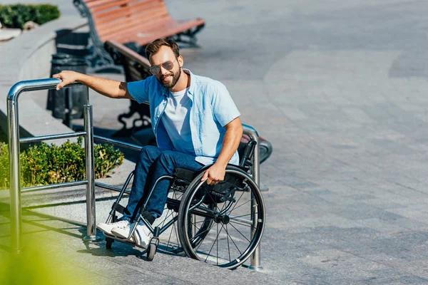 Hombre guapo en gafas de sol usando silla de ruedas en escaleras sin rampa y mirando a la cámara - foto de stock