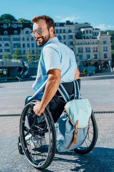 Sorrindo homem bonito em óculos de sol usando cadeira de rodas com saco na rua e olhando para longe — Fotografia de Stock