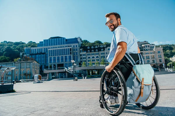 Smiling handsome man in sunglasses using wheelchair on street and looking at camera — Stock Photo