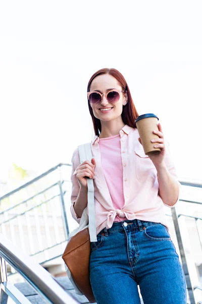 Attractive redhair woman in pink shirt looking at camera on stairs with disposable coffee cup — Stock Photo