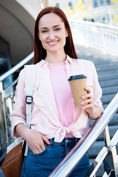 Portrait of attractive redhair woman in pink shirt with disposable coffee cup looking at camera on stairs — Stock Photo
