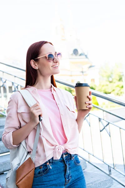 Sonriente atractiva pelirroja mujer en camisa rosa sosteniendo café en taza de papel y mirando hacia otro lado - foto de stock