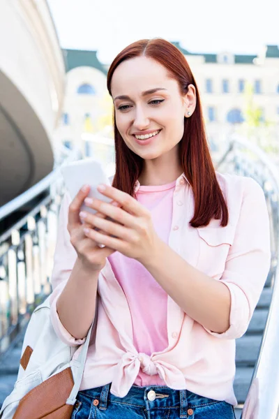 Enfoque selectivo de sonriente mujer pelirroja atractiva en camisa rosa usando teléfono inteligente en las escaleras - foto de stock