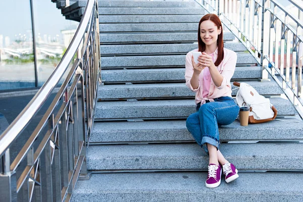 Smiling attractive redhair woman in pink shirt sitting on stairs and using smartphone — Stock Photo
