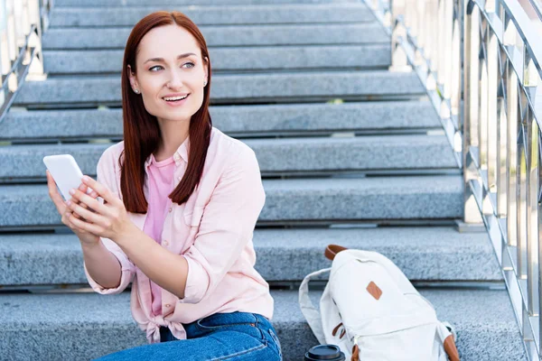 Sorridente donna capelli rossi attraente in camicia rosa seduto sulle scale e utilizzando smartphone, guardando altrove — Foto stock