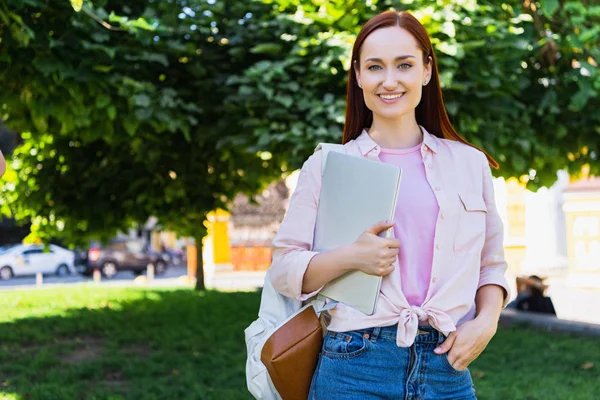 Atractivo freelancer alegre sosteniendo portátil y mirando a la cámara en el parque - foto de stock