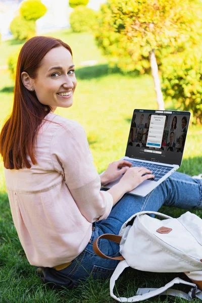 Sonriente atractiva mujer usando el ordenador portátil con página linkedin cargado en el parque y mirando a la cámara - foto de stock