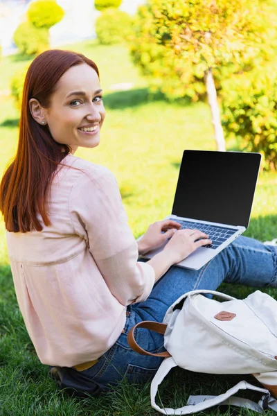 Sonriente freelancer atractivo usando portátil con pantalla en blanco en el parque y mirando a la cámara - foto de stock