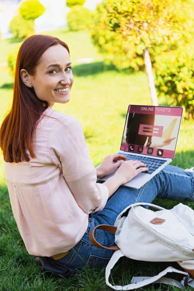 Улыбающаяся привлекательная женщина с помощью ноутбука с онлайн-билетов прибор в парке и смотреть в камеру — стоковое фото