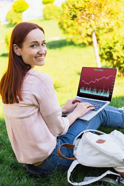 Smiling attractive woman using laptop with traiding appliance in park and looking at camera — Stock Photo