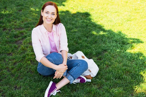 Sonriente atractiva mujer sentado en verde hierba en el parque y mirando a la cámara - foto de stock