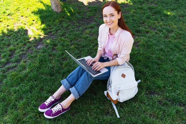 Vue grand angle de sourire attrayant pigiste en utilisant un ordinateur portable dans le parc et en regardant la caméra — Photo de stock