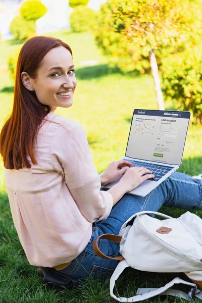 Souriant femme attrayante en utilisant un ordinateur portable avec la page facebook chargée dans le parc et en regardant la caméra — Photo de stock