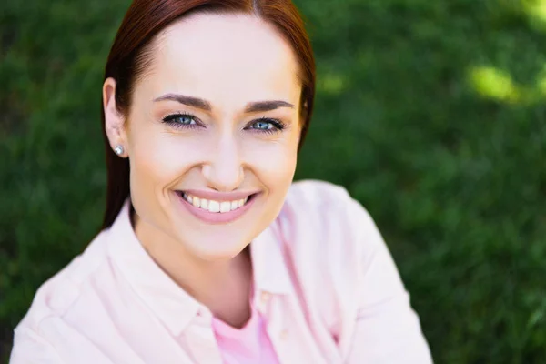 Tête et les épaules de belle femme rousse heureuse en chemise rose regardant la caméra dans le parc — Photo de stock