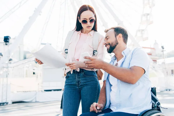 Handsome boyfriend in wheelchair using smartphone and girlfriend standing with map on street — Stock Photo