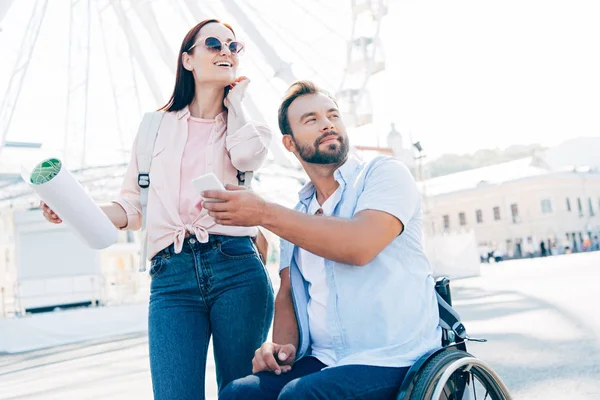 Красивый парень в инвалидной коляске показывает смартфон подружке с картой на улице — стоковое фото