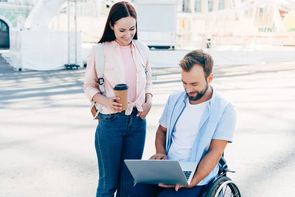 Улыбающийся красивый парень в инвалидной коляске с помощью ноутбука и подружки, стоящих с кофе в бумажной чашке на улице — стоковое фото