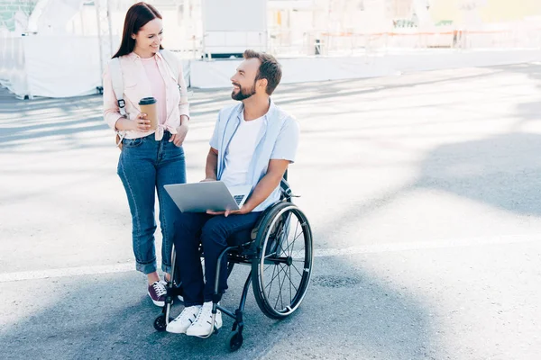 Guapo novio en silla de ruedas usando portátil y novia de pie con café en taza de papel en la calle, mirándose el uno al otro - foto de stock