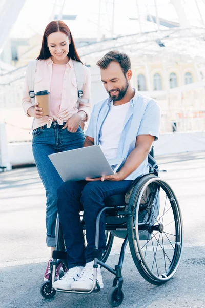 Guapo novio en silla de ruedas usando portátil y novia de pie con café para ir en la calle - foto de stock