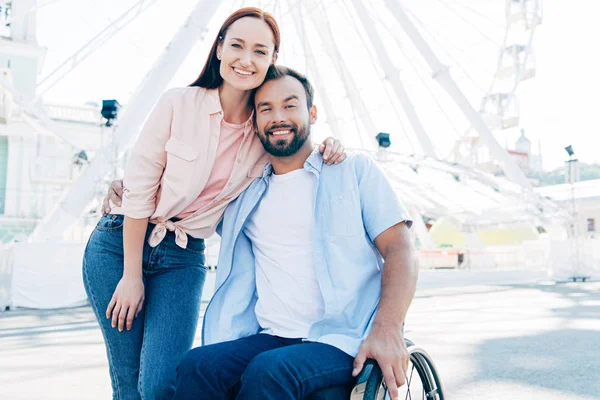 Smiling handsome boyfriend in wheelchair and girlfriend hugging and looking at camera on street near observation wheel — Stock Photo