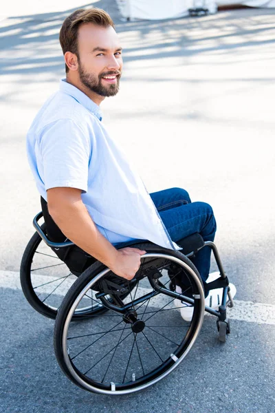 Handsome smiling man using wheelchair on street and looking at camera — Stock Photo