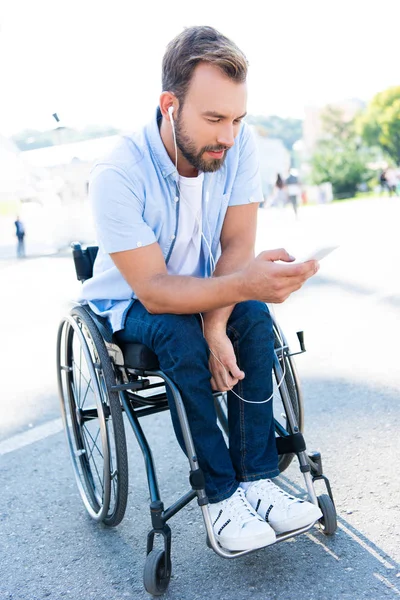 Hombre guapo en silla de ruedas escuchando música con teléfono inteligente en la calle - foto de stock