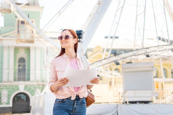 Attractive smiling tourist in sunglasses standing with map on street and looking away — Stock Photo