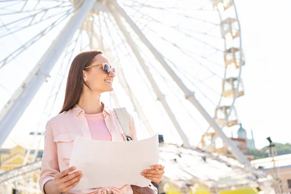 Smiling attractive tourist in sunglasses standing with map near observation wheel and looking away — Stock Photo