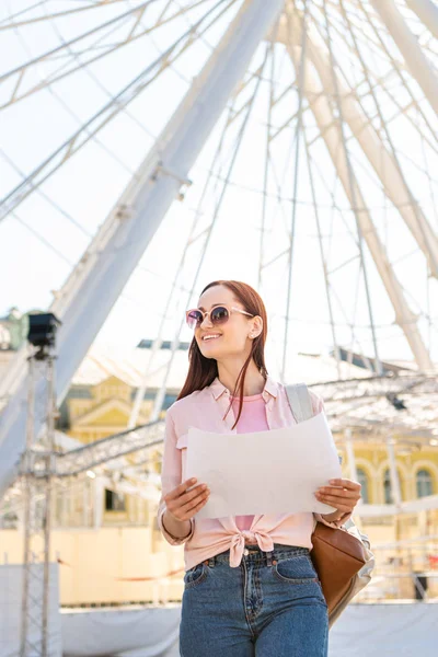 Attraente turista sorridente in occhiali da sole in piedi con mappa vicino alla ruota panoramica nel parco divertimenti — Foto stock