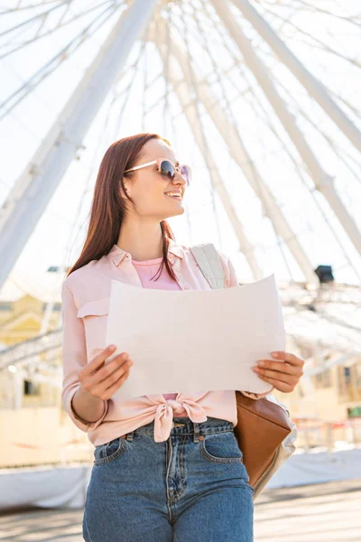 Attraente turista sorridente in occhiali da sole in piedi con mappa vicino alla ruota panoramica nel parco divertimenti e guardando altrove — Stock Photo