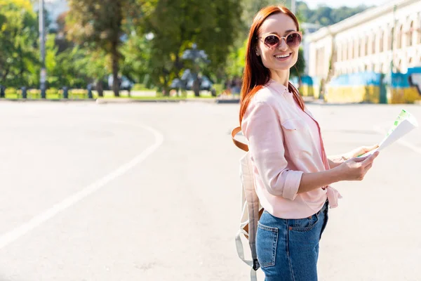 Turista atractivo feliz en gafas de sol de pie con mapa en la calle y mirando a la cámara - foto de stock