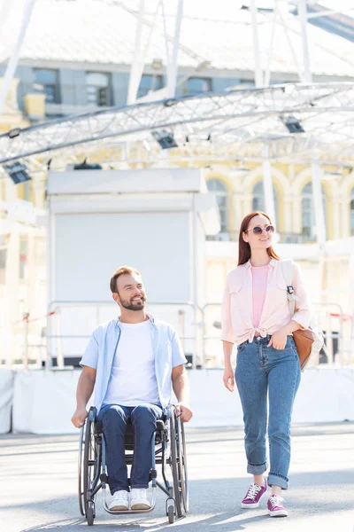 Красивый парень в инвалидной коляске и привлекательная девушка, идущая по улице — стоковое фото