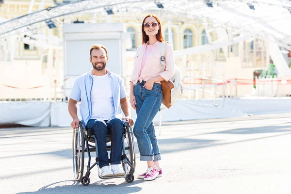 Heureux beau petit ami en fauteuil roulant et petite amie regardant caméra sur la rue — Photo de stock