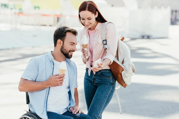 Красивый парень в инвалидной коляске и девушка с мороженым, глядя на смартфон на улице — стоковое фото