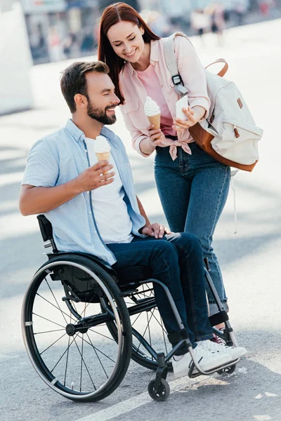 Girlfriend with ice cream showing something on smartphone to handsome boyfriend in wheelchair on street — Stock Photo