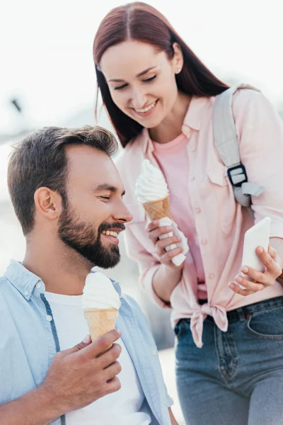 Retrato de novia con helado mostrando algo en el teléfono inteligente a novio en silla de ruedas en la calle - foto de stock