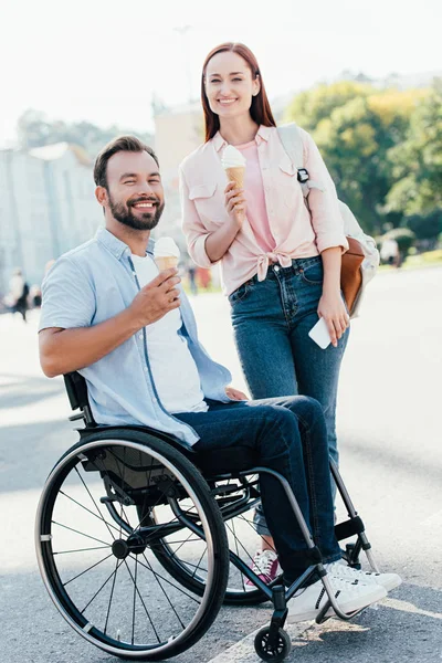 Sourire beau petit ami en fauteuil roulant et petite amie avec de la crème glacée regardant caméra sur la rue — Photo de stock