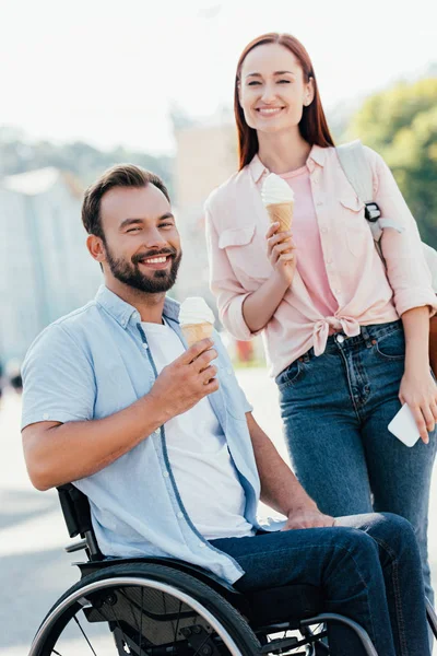 Happy handsome boyfriend in wheelchair and girlfriend with ice cream looking at camera on street — Stock Photo