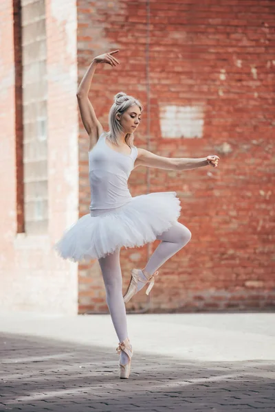 Attractive young ballerina in white tutu and pointe shoes dancing on street — Stock Photo
