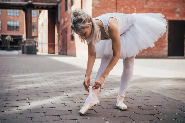 Young ballerina in white tutu skirt tying pointe shoe on street — Stock Photo