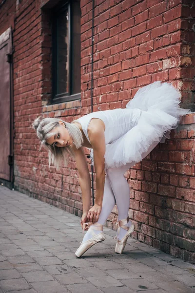 Attractive young ballerina leaning at brick wall and tying pointe shoe — Stock Photo