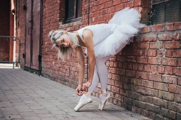 Beautiful young ballerina leaning at brick wall and tying pointe shoe — Stock Photo