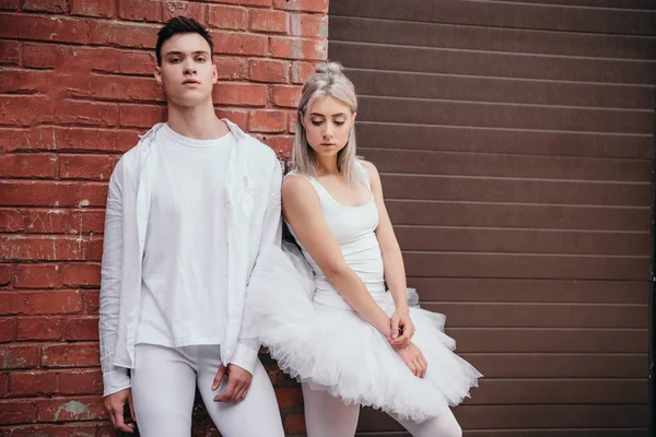 Pareja de jóvenes bailarines en ropa blanca de pie juntos cerca de la pared de ladrillo en la calle - foto de stock