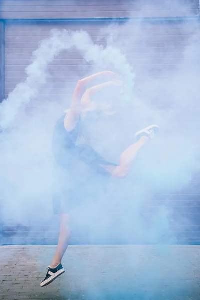 Молодая современная танцовщица танцует в голубом дыму на улице — стоковое фото
