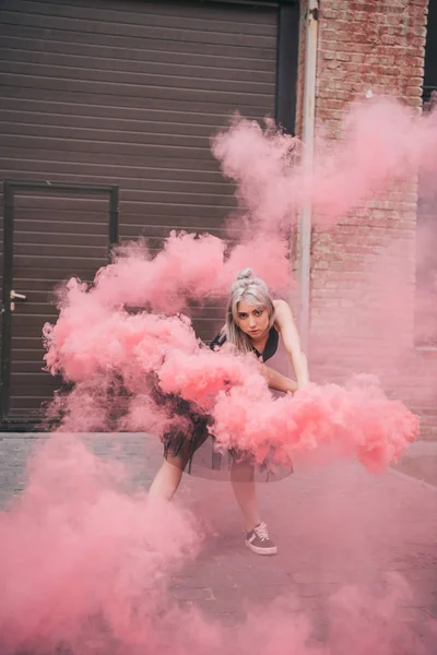 Attraente giovane donna che balla in rosa fumo e guardando la fotocamera sulla strada — Foto stock