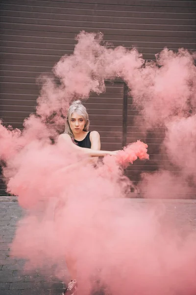 Giovane donna che guarda la macchina fotografica mentre balla in fumo rosa sulla strada — Foto stock