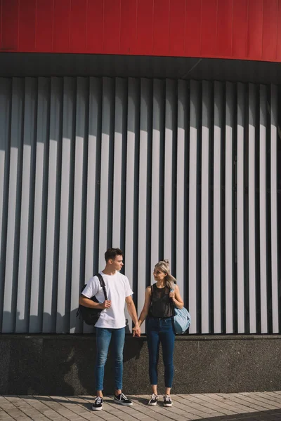 Beautiful stylish young couple with backpacks holding hands and looking at each other on street — Stock Photo