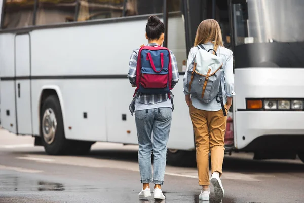 Rear view of female tourists with backpacks walking near travel bus at urban street — Stock Photo