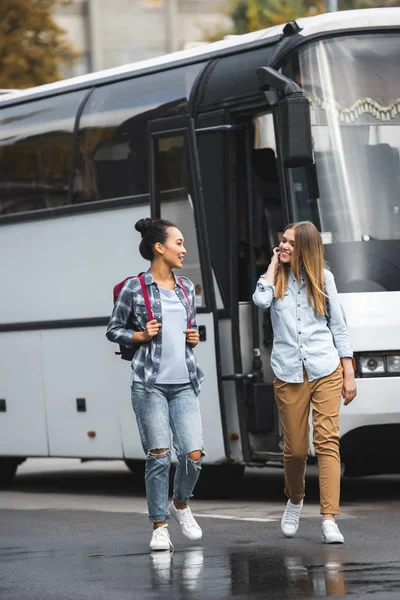 Selective focus of multicultural women with backpacks walking near travel bus at urban street — Stock Photo